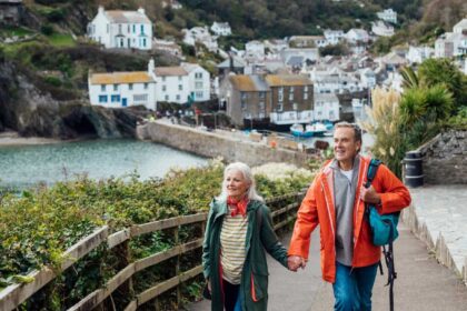 A senior man and his wife holding hands walking up a hill on a footpath looking away from the camera at the view. The fishing village of Polperro is behind them.