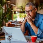 Middle-aged white man wearing glasses, staring into space over the top of his laptop in a coffee shop