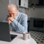 Portrait of pensive bearded senior looking on screen of laptop sitting at table with coffee cup.