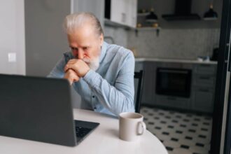 Portrait of pensive bearded senior looking on screen of laptop sitting at table with coffee cup.