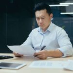 Asian man looking concerned while studying paperwork at his desk in an office